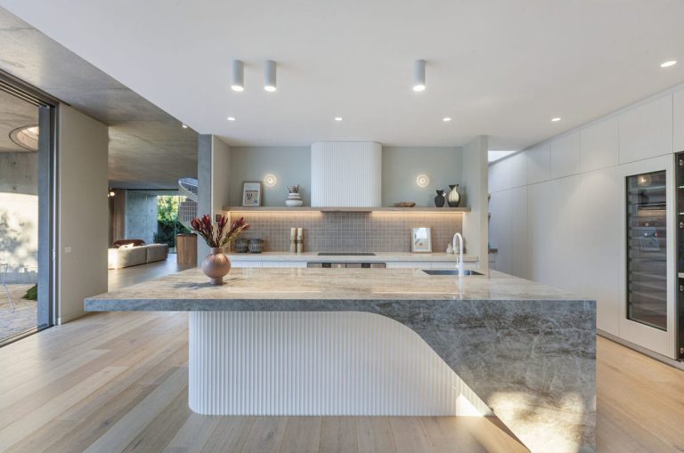 A contemporary kitchen featuring light grey cabinetry, a marble countertop and backsplash, built-in stainless steel appliances, and modern pendant lighting, with natural light streaming in through large windows.